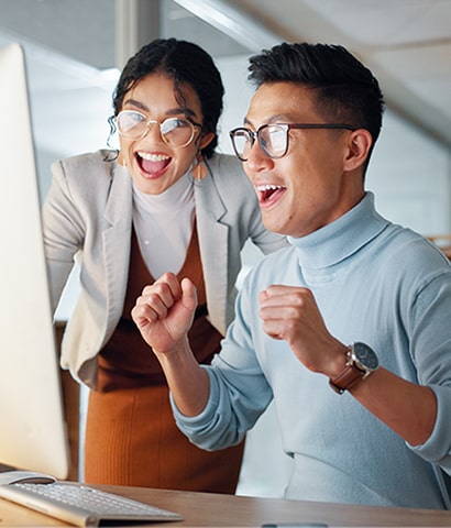 Two excited coworkers smiling and cheering as they look at a computer screen in an office setting.